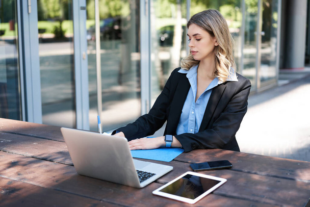 Porträt einer Frau, die an der frischen Luft arbeitet, mit Laptop, digitalem Tablet auf einer Bank sitzt und Notizen macht. - Foto, Bild