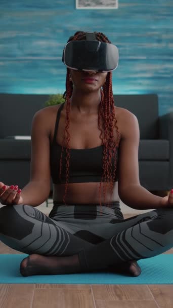 Verical video: Fit μαύρη γυναίκα που φοράει ακουστικά εικονικής πραγματικότητας ενώ κάθεται στο χάρτη της γιόγκα σε στάση λωτού κατά τη διάρκεια ασκήσεων διαλογισμού στο σαλόνι. Αξιολύπητος ενήλικας απολαμβάνοντας υγιή τρόπο ζωής - Πλάνα, βίντεο