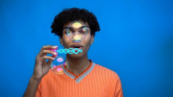 Junger afrikanisch-amerikanischer Mann bläst Seifenblasen und schaut isoliert in die Kamera auf blauem Grund  - Foto, Bild
