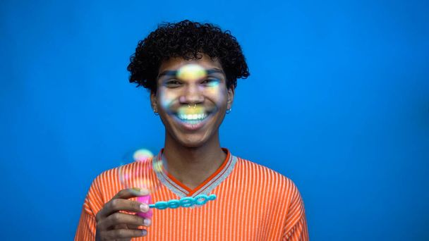 Χαμογελώντας Αφροαμερικάνος κρατώντας ραβδί κοντά σε σαπουνόφουσκες απομονωμένες στο μπλε  - Φωτογραφία, εικόνα