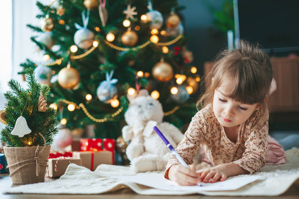 Carino bambina scrivere lettera a Babbo Natale o scrivere sogni di un regalo con vicino albero di Natale. Buon Natale e Felice Anno Nuovo! - Foto, immagini