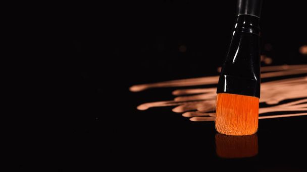 Vista de cerca del cepillo cosmético cerca de la base de maquillaje borrosa sobre fondo negro  - Foto, imagen