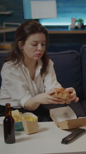 Pionowe wideo: Biała kobieta jedząca hamburgera z torby dostawczej podczas oglądania filmu w domu. Dorosły korzystających na wynos fast food posiłek i butelka piwa siedzi na kanapie w salonie - Materiał filmowy, wideo