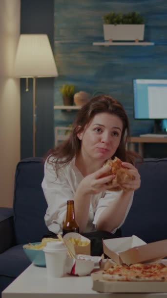 Pionowe wideo: Osoba jedząca pyszne hamburgery i frytki w salonie podczas picia napojów alkoholowych i oglądania telewizji. Kobieta z jedzeniem na wynos fast food posiłek siedzi na kanapie i relaksujące - Materiał filmowy, wideo