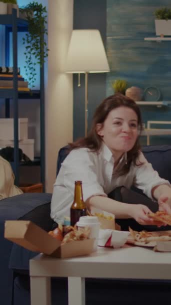 Pionowe wideo: Osoba jedząca kawałek pizzy podczas oglądania komedii w telewizji i śmiejąca się na kanapie w domu. Biała kobieta zabawia się filmem w telewizji, delektując się fast foodem na wynos i butelką - Materiał filmowy, wideo