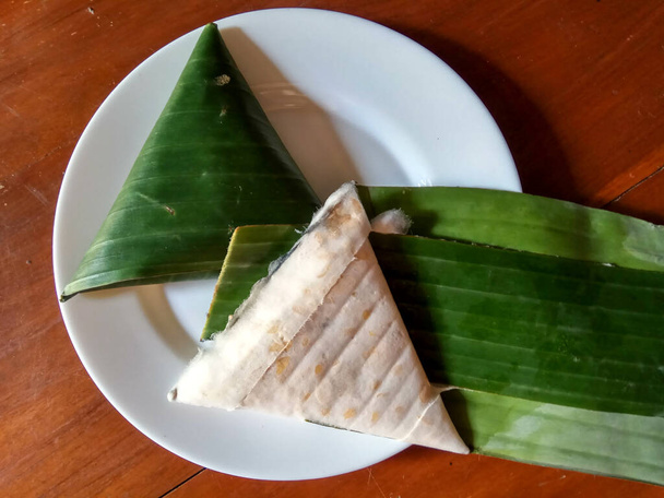 Tempeh ou tempe, produto de soja processado, envolto em folhas de banana com forma triangular. Colocado em placa branca, com fundo de madeira - Foto, Imagem