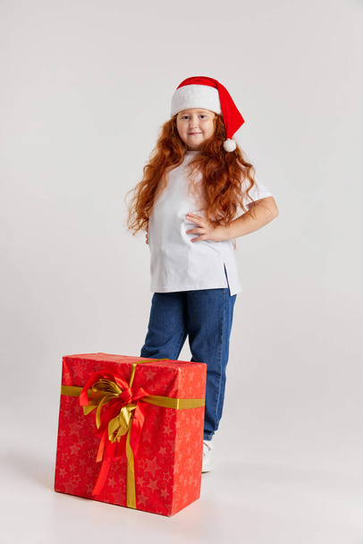 Happy smiling child in casual outfit and Christmas hat with boxes in festive package. Girl rejoices at holiday. Merry Christmas, New Year. Concept of emotions, dreams, happy childhood, wishes - Photo, image