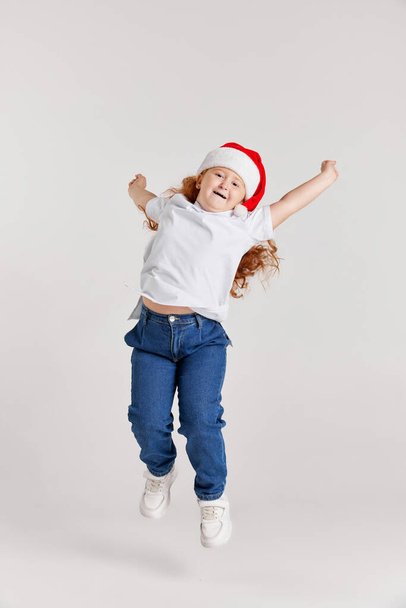 Christmas mood. Thrilled school age girl, kid in white t-shirt and jeans jumping high isolated over white background. Kids fashion, emotions, carefree childhood, challenges, education concept - Foto, imagen
