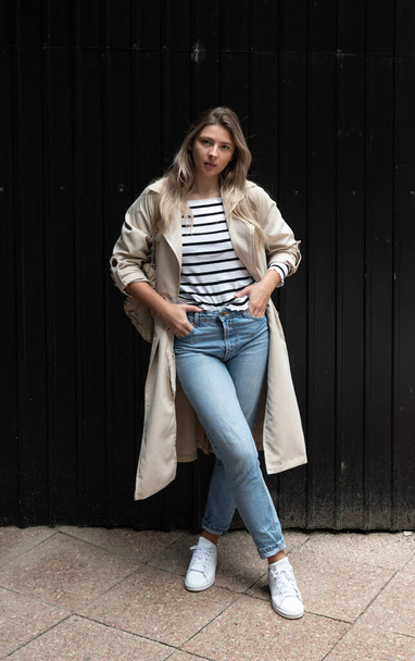 Portrait of attractive lady in checkered blue jeans and beige trench coat looks and poses outside. Fashionable woman outdoor posing in cozy stylish outfit - 写真・画像