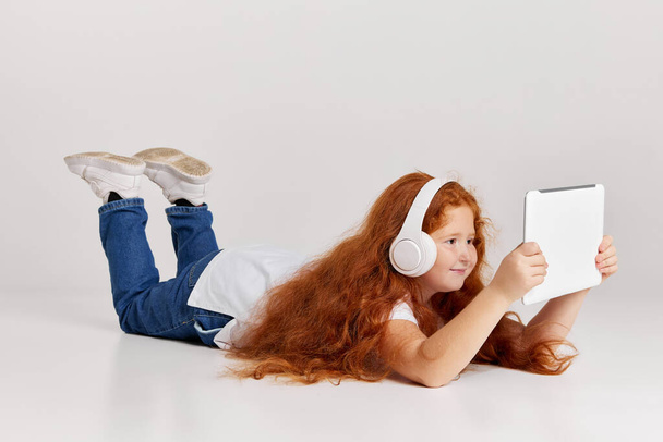 Watching cartoons. Cute redheaded little girl wearing tee and jeans using tablet isolated over white studio background. Remote online education, kids emotion, facial expressions, fashion - Фото, зображення