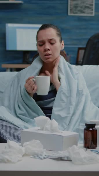 Vertical video: Woman with seasonal cold holding cup of tea wrapped in blanket. Sick adult with flu looking at camera while feeling ill and shivering. Portrait of person with medicaments on table - Footage, Video