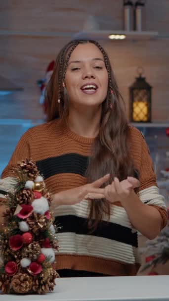 垂直ビデオ:クリスマスシーズンに装飾されたお祝いのキッチンでビデオ通話カメラで手を振る若い大人。冬の装飾品や装飾品と部屋に座っている間に茶を準備セーターを持つ女性 - 映像、動画