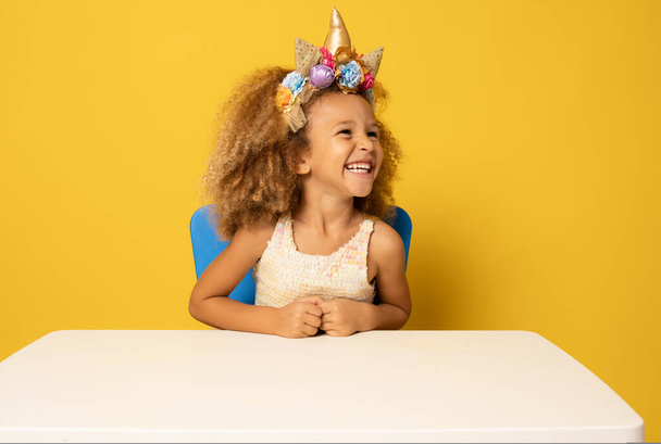 Roztomilé dítě dívka v jednorožec čelenka slaví narozeninovou oslavu přes žluté pozadí. - Fotografie, Obrázek