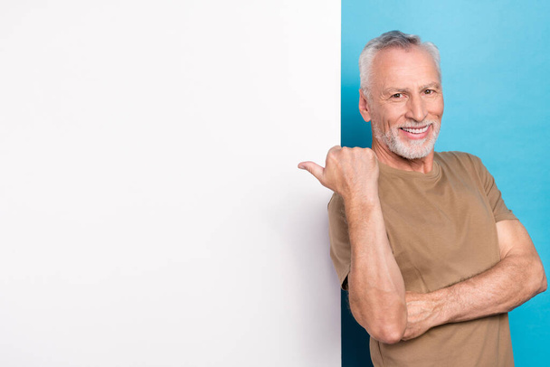 Portrait of positive cheerful retired man with gray hair beard beige t-shirt directing empty space isolated on blue color background. - Фото, изображение