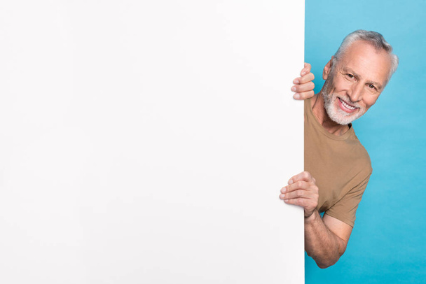 Portrait of optimistic funny retired man with gray hair beard beige t-shirt look out of white placard isolated on blue color background. - 写真・画像
