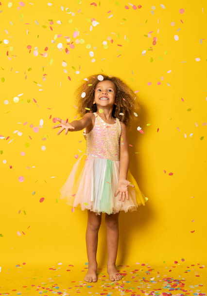 Lustiges kleines Mädchen für die Party in Pailletten und Tutu Konfetti blasen gekleidet. Vereinzelt auf gelbem Hintergrund. - Foto, Bild