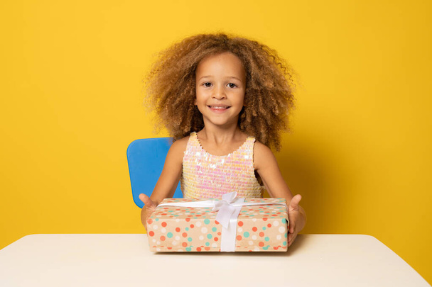 Cute child girl in party dress holding gift box isolated over yellow background. - Photo, Image