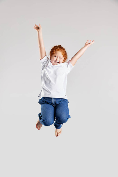 Happy holidays. Thrilled school age girl, kid in white t-shirt and jeans jumping high isolated over white background. Kids fashion, emotions, carefree childhood, challenges, education concept - Фото, зображення