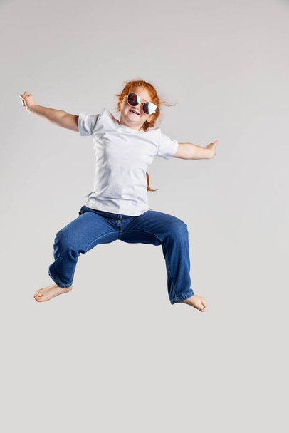 Dance. Joyful little girl, kid in white t-shirt and jeans jumping high isolated over white background. Kids fashion, emotions, carefree childhood, challenges, education concept - Fotografie, Obrázek