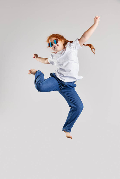 Bav se. Šťastná holčička, dítě v bílém tričku a džínách skákající vysoko izolovaně přes bílé pozadí. Dětská móda, emoce, bezstarostné dětství, výzvy, koncepce vzdělávání - Fotografie, Obrázek