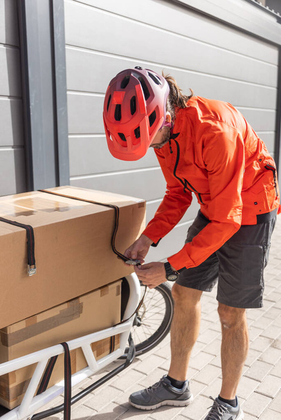 赤い服を着た若い宅配便と貨物自転車に乗るヘルメット市の住所に荷物を配達するために目的地に到着貨物を積み降ろし - 写真・画像