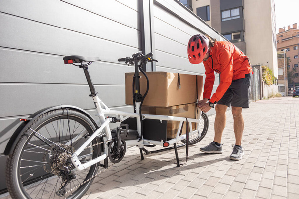 young courier with red clothing and helmet riding cargo bike arriving at the shipping destination to deliver a package to a city address, unloading the cargo - Photo, Image