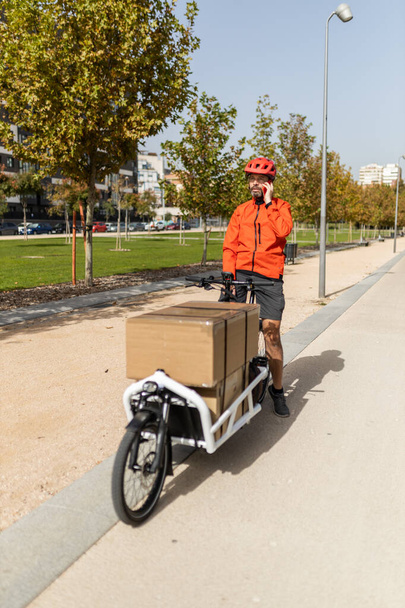 young courier with red clothing and helmet riding cargo bike , riding along the city bike path on his way to deliver a package, talking on the phone with the customer - Foto, afbeelding