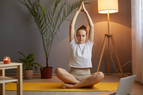 Indoor shot of beautiful woman with hair bun wearing white t shirt practicing yoga at home while sitting on floor on mat, raised her arms, looking at camera with serious expression. - Foto, afbeelding