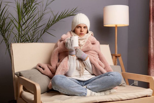 Image of young adult woman wearing warm clothes and wrapped in scarf sitting on sofa with cup of tea in hands, trying to get warm, drinking hot beverage. - 写真・画像