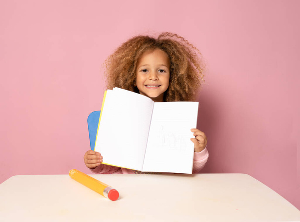 Education. Close up portrait of cute child little girl is sitting at the table with a notebook for drawing, isolated on pink background - Foto, Bild