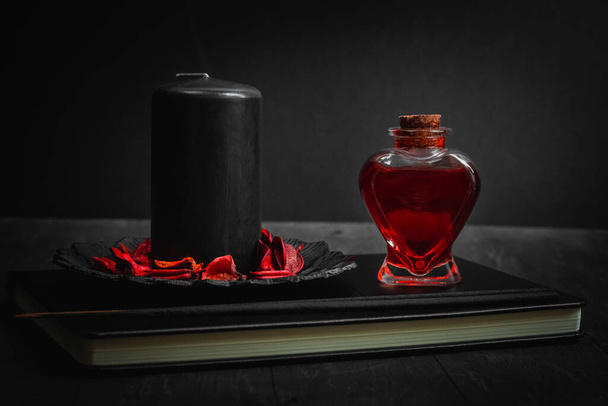 A black candle with a red potion in a heart shape bottle and ritual sticks lie on a magic book in the center on a black wooden background, close-up side view. Esoteric concept, dark style. - Foto, Bild