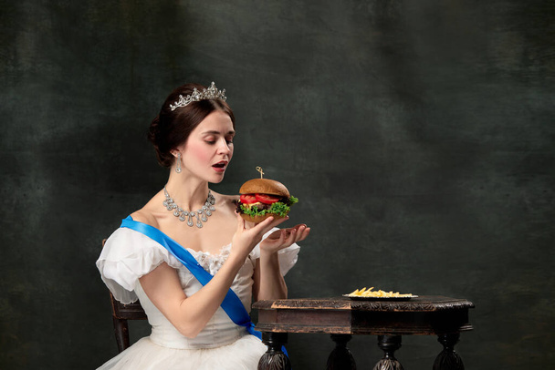 Wow. Young beautiful woman, royal person, queen or princess in white medieval outfit tasting burger on dark background. Concept of comparison of eras, modern, fashion, beauty. - Photo, image