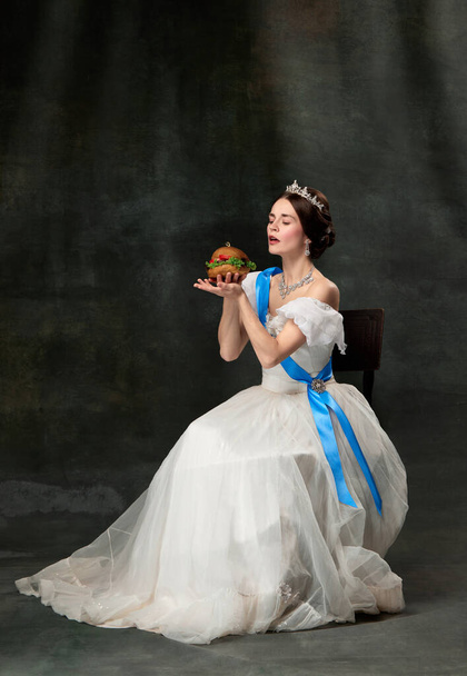 Desire. Elegant woman, royal person, queen or princess in white medieval outfit tasting juicy hamburger on dark vintage background. Concept of comparison of eras, modern, fashion, beauty. - Foto, imagen