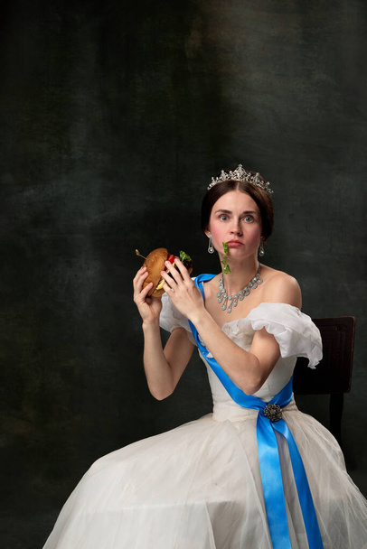 Fast food or dieting. Young funny girl like queen or princess in white medieval outfit eating burger on dark background. Concept of comparison of eras, modern, fashion, beauty and ad - Foto, imagen