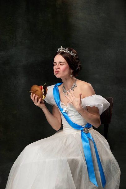 Enjoy the taste. Beautiful young queen or princess in white medieval outfit eating burger on dark vintage background. Concept of fast food, comparison of eras, modern vision, fashion, beauty. - Foto, imagen