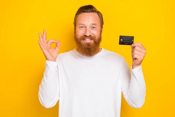 Photo of positive stylish man trendy outfit get arm hold plastic bank card show okey symbol recommend isolated on yellow color background. - Фото, изображение