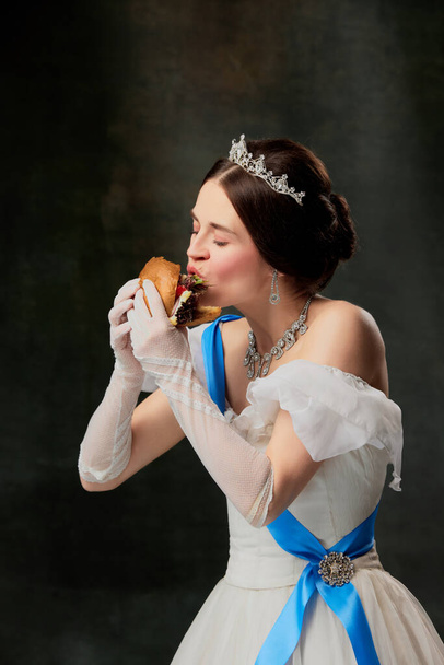 Enjoy the taste. Beautiful young queen or princess in white medieval outfit eating burger on dark vintage background. Concept of fast food, comparison of eras, modern vision, fashion, beauty. - Photo, image