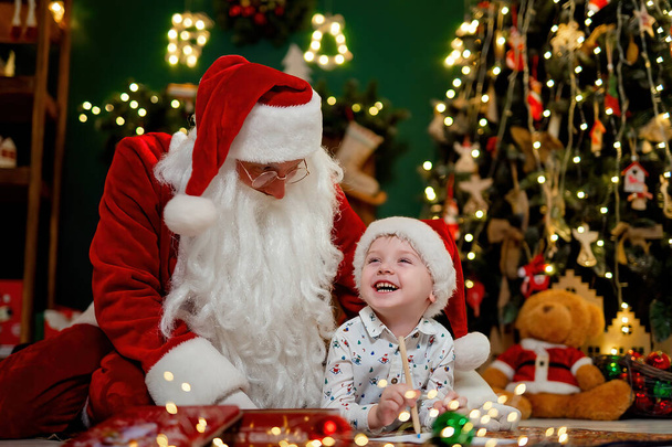 Little cute boy in Santas hat having fun time with Santa Claus while lying on the floor near Christmas tree. New Year concept  - Фото, изображение