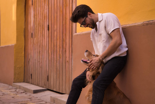 Νεαρός Ισπανός με γενειάδα, γυαλιά ηλίου και λευκό πουκάμισο, ακουμπισμένος στον τοίχο αγκαλιάζοντας το σκύλο του ανάμεσα στα πόδια του. Έννοια ζώα, τα σκυλιά, την αγάπη, τα κατοικίδια ζώα, χρυσό. - Φωτογραφία, εικόνα