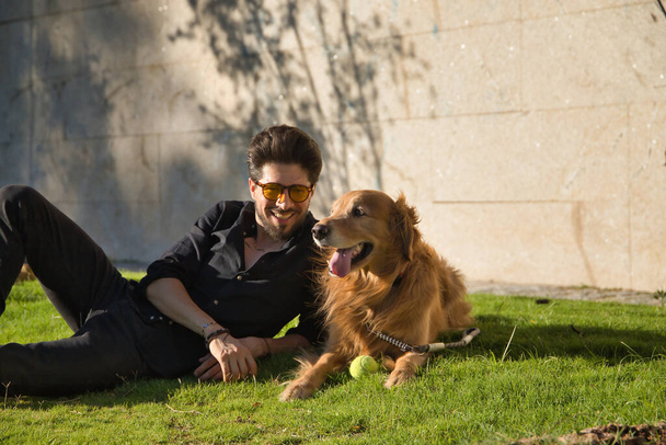 Young Hispanic man with a beard, sunglasses and black shirt, lying on the lawn next to his dog sunbathing. Concept animals, dogs, love, pets, gold. - Foto, afbeelding