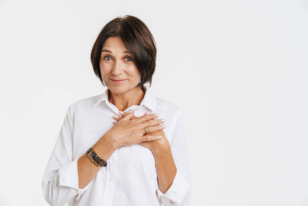 Mature brunette woman smiling and holding hands on her chest isolated over white background - Photo, image