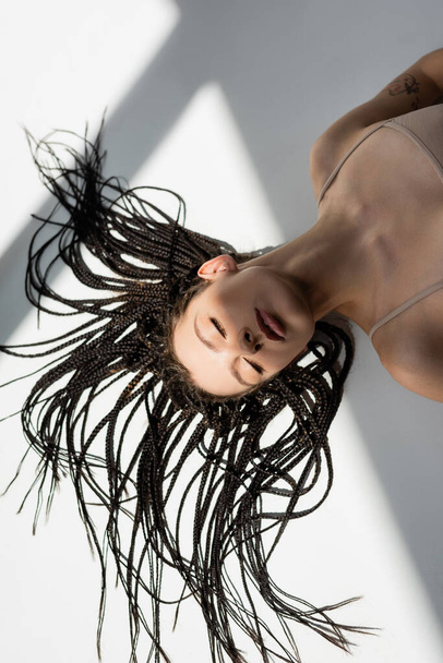 Visão superior da jovem com tranças deitadas sobre fundo cinza  - Foto, Imagem