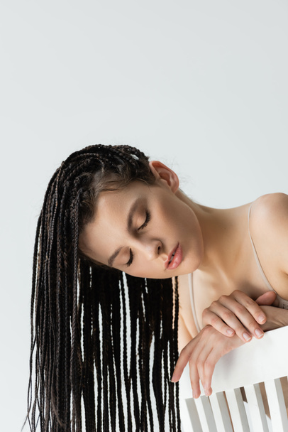Young woman with braids posing with closed eyes near chair isolated on grey  - Foto, Imagem