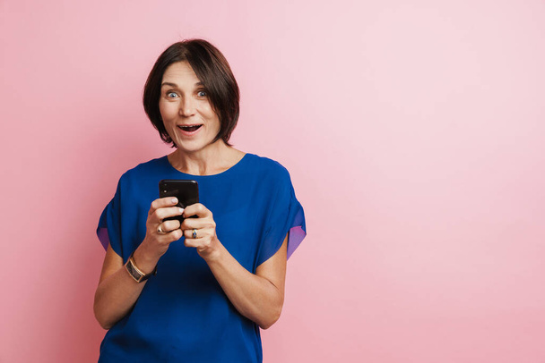 Mature woman expressing surprise and using mobile phone isolated over pink background - Valokuva, kuva