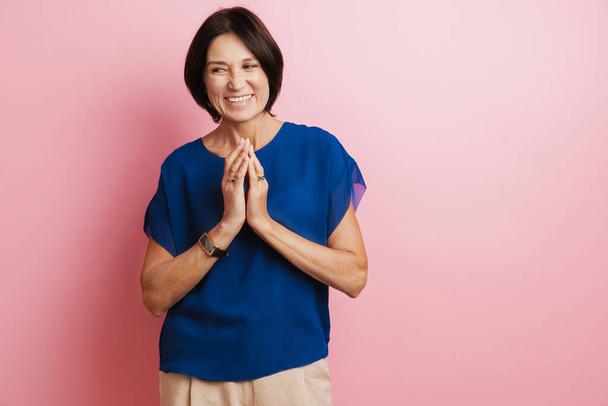 Mature brunette woman laughing while holding hands together isolated over pink background - Photo, image