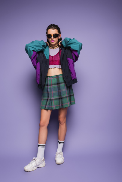 Trendy young model in plaid skirt and sports jacket standing on purple background  - Foto, Imagem