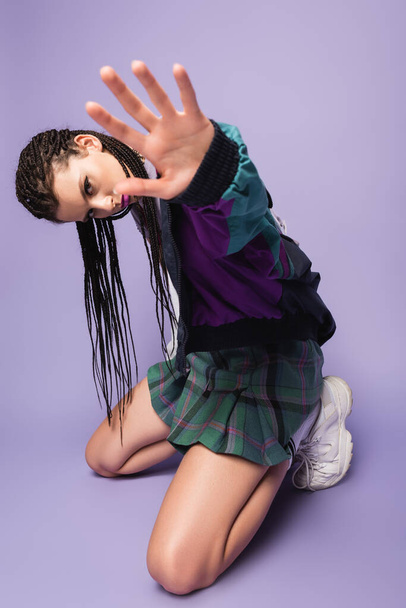 stylish woman with braided dreadlocks kneeling in vintage clothes with outstretched hand on purple background - Photo, Image