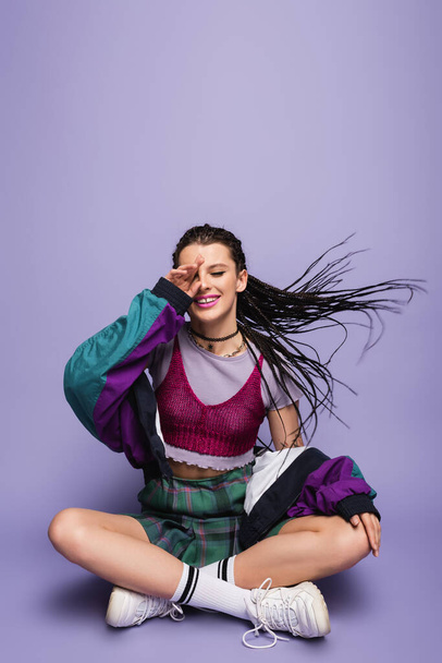 smiling woman in nineties attire sitting with crossed legs on wind on purple background - Foto, Imagem