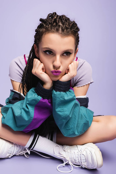 thoughtful nineties style woman sitting with crossed legs and pouting lips on purple background - Fotó, kép