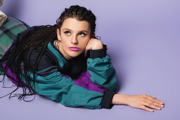 dreamy woman with braids lying in vintage style jacket and looking away on purple background - Foto, Imagen